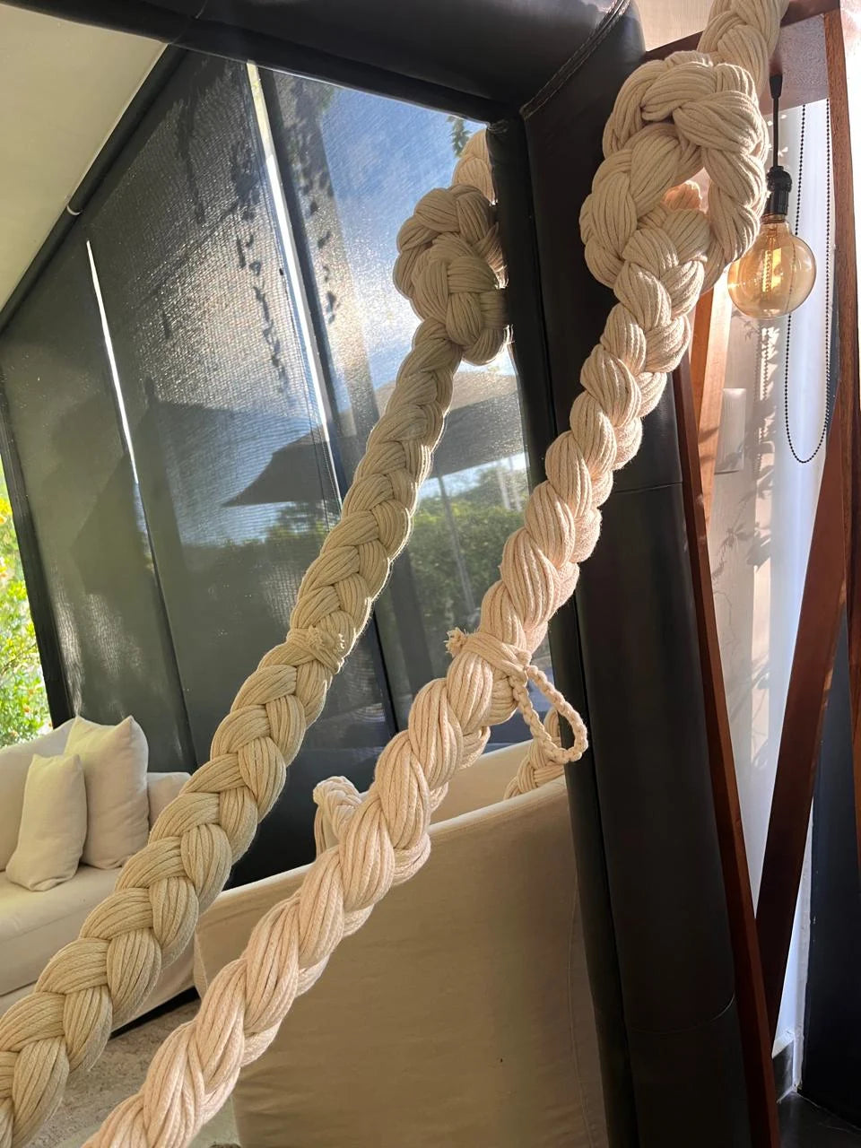 Pale braids suspended diagonally in front of a window beside white armchairs.