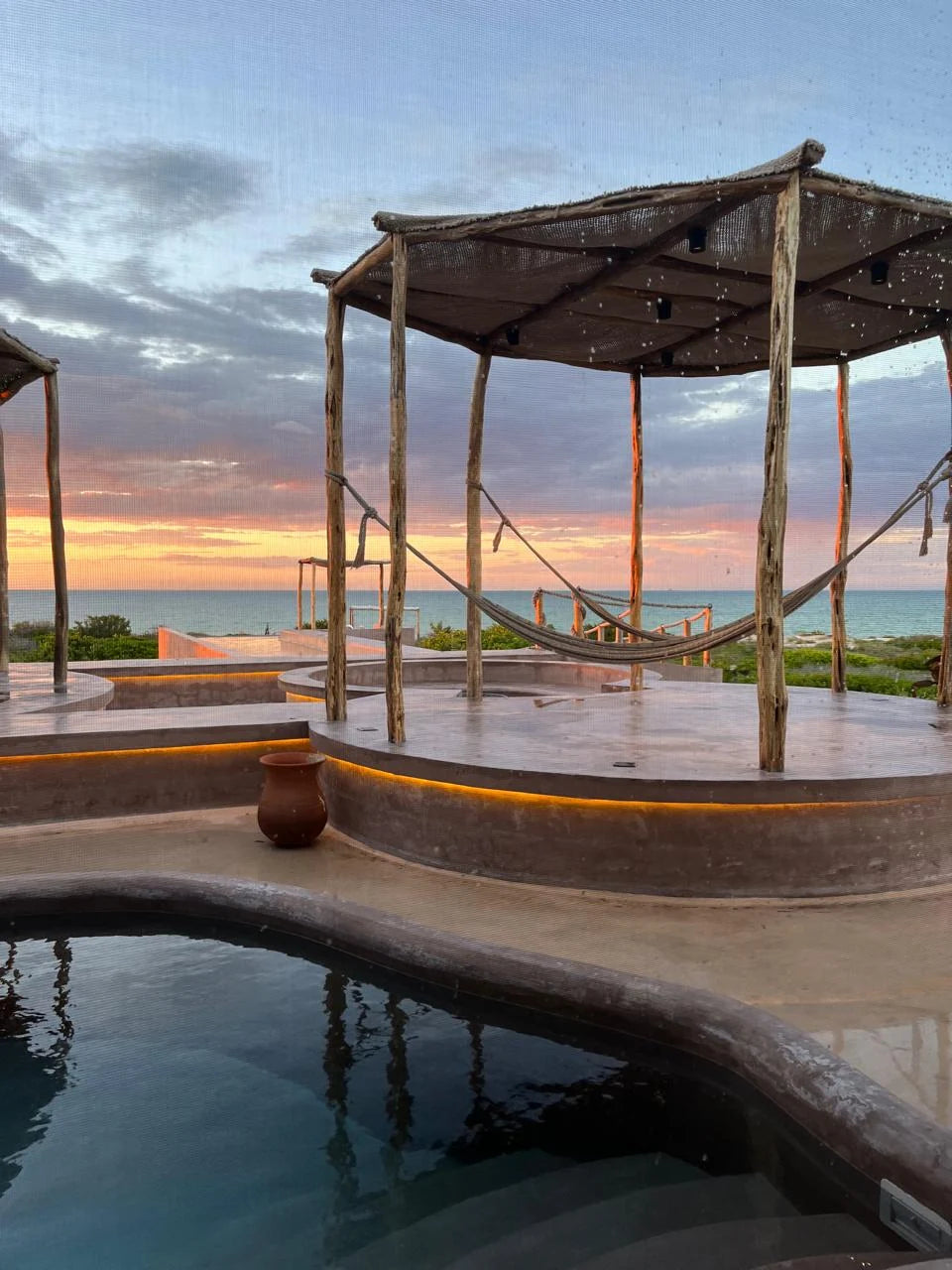 Hammocks beneath a wooden pavilion beside a circular pool at sunset.