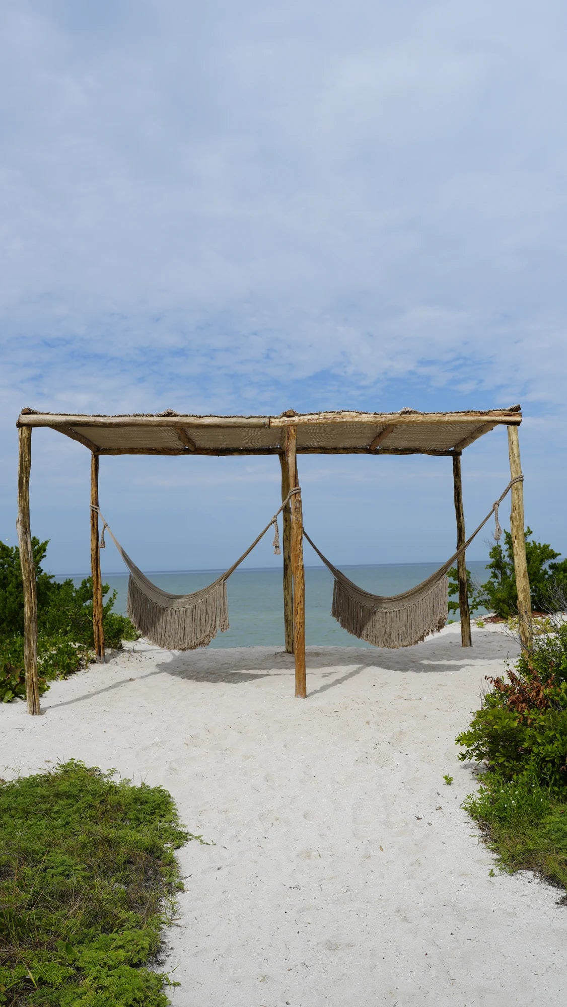 Two hammocks beneath a woven canopy facing the sea, surrounded by sand and greenery.