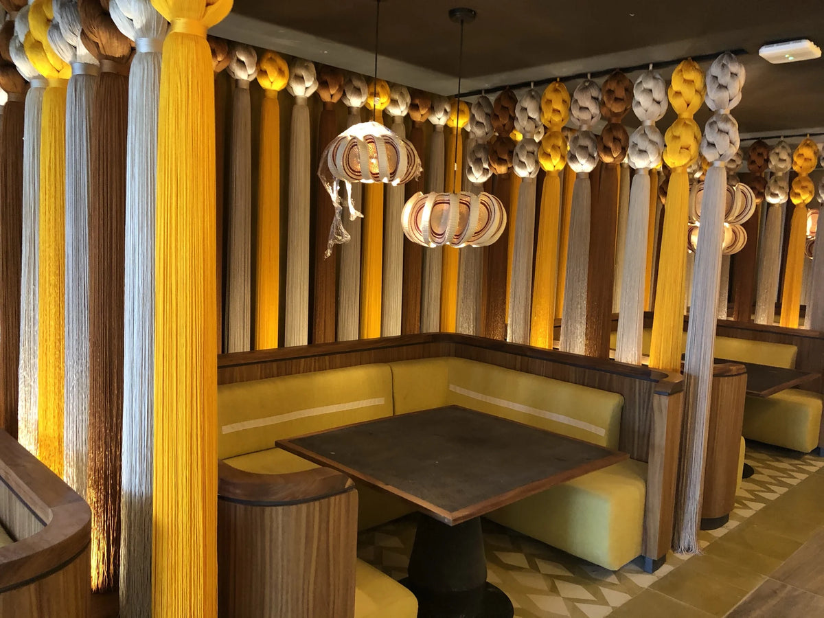 A table and banquette seating framed by vertical threads and hanging lights at Kibi Kibi.