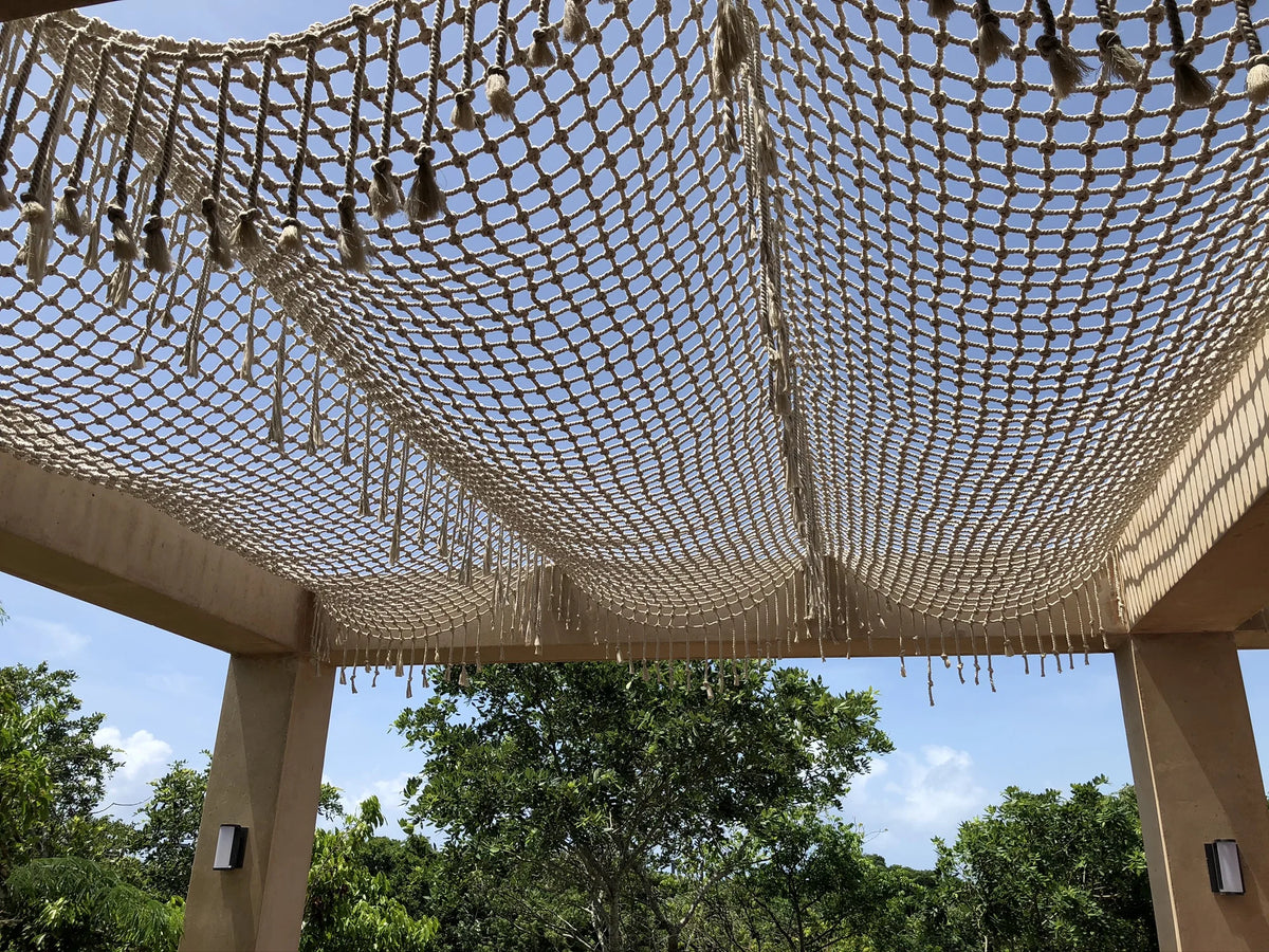 Detail of the knotted canopy and fringe above the pergola’s pillars, with blue sky and trees beyond.